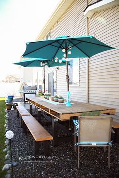 an outdoor table with two umbrellas over it and some chairs around it on gravel