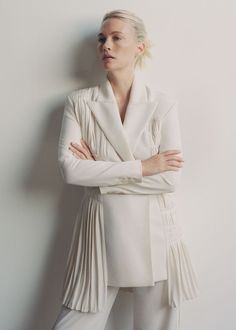 a woman standing with her arms crossed wearing a white jacket and pants, leaning against a wall