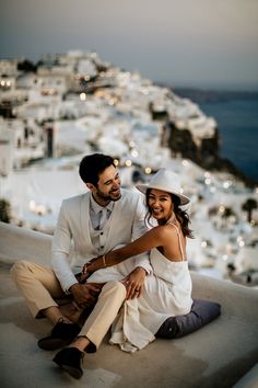 a man and woman sitting on top of a hill next to the ocean with buildings in the background