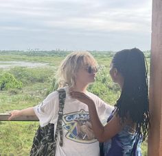 two women standing next to each other looking at the grass and trees in the distance