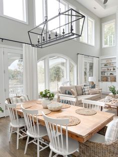 a dining room table surrounded by chairs and white couches with windows in the background