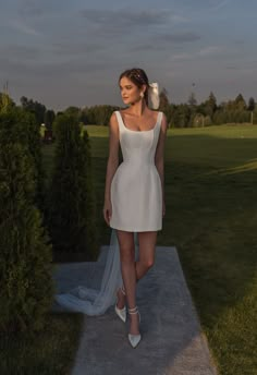 a woman in a short white dress is standing on a sidewalk near bushes and trees