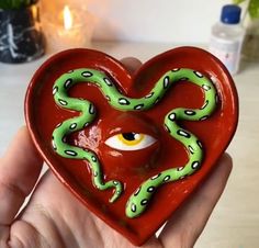 a hand holding a red heart with a green snake on it and an eye in the center