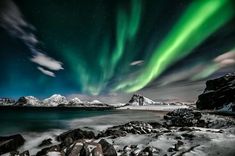 the northern lights shine brightly over snow covered rocks and water in front of snowy mountains