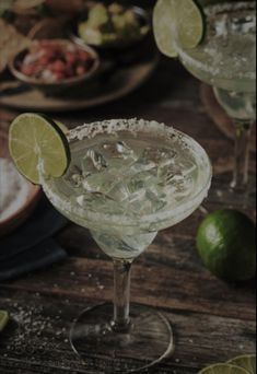 two margaritas sitting on top of a wooden table next to lime wedges and chips