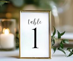 a table number is displayed in a gold frame with greenery and candles behind it