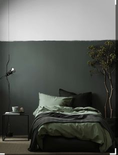 a bed with green sheets and pillows next to a small potted tree in the corner