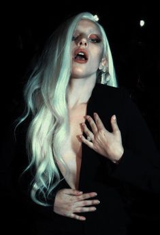 a woman with white hair and makeup is posing for the camera in black dress, holding her hands on her chest