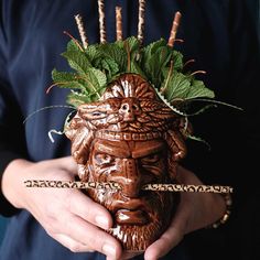 a person holding a mask with green leaves on it's head and sticks in their hands