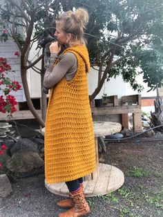 a woman standing in front of a tree wearing a yellow crochet vest and brown boots