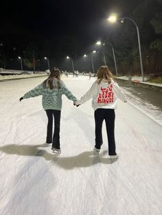 two girls are holding hands while skating on an ice rink at night with street lights in the background