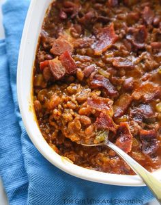 a white bowl filled with beans and bacon on top of a blue towel next to a spoon