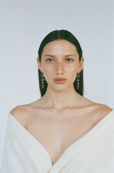 a woman with long dark hair in a white top and earrings on her head, looking at the camera