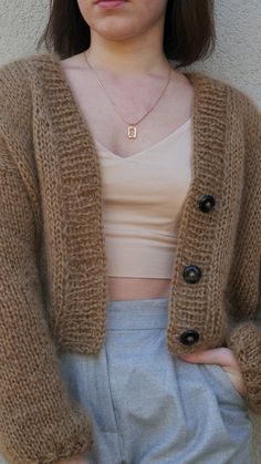 a woman standing in front of a white wall wearing a brown cardigan sweater and light blue shorts