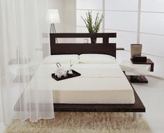 a bed with white sheets and pillows in a room next to a plant on a table