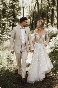 a bride and groom are walking through the woods in their wedding attire, holding hands