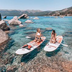 two women are sitting on surfboards in the clear blue water near rocks and boulders