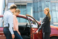 two women and a man are shaking hands in front of a red car with the door open