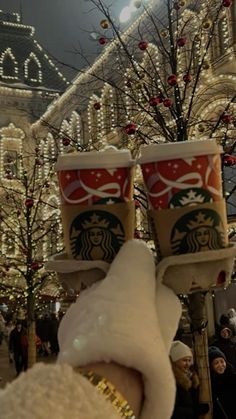 two starbucks cups are held up in front of a christmas tree and decorated with lights