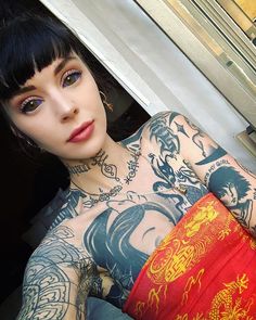 a woman with tattoos on her arm and chest sitting in front of a window, looking at the camera