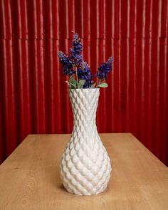 a white vase filled with purple flowers on top of a wooden table next to a red wall