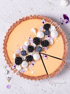 a cheesecake topped with berries and blackberries on top of a white table next to purple flowers