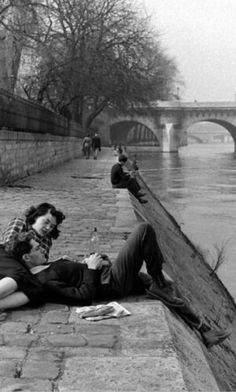 black and white photograph of two people laying on the ground next to water with bridge in background