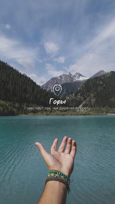 a person's hand reaching out to the water with mountains in the background and blue sky above