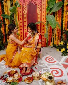 two women in yellow dresses sitting on the ground next to some plates and bowls filled with food