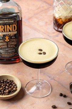 two glasses filled with coffee sit on a table next to a bottle and some beans