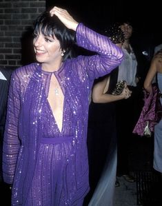 a woman in a purple dress dancing with her hands behind her head and smiling at the camera