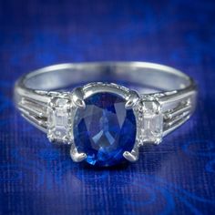 An elegant Edwardian inspired trilogy ring adorned with a deep, royal blue sapphire in the centre weighing 1.65ct. It's complemented beautifully by two bright white, emerald cut diamonds at each side with exceptional VS 1 clarity - H colour (approx. 0.16ct each). The stones are claw set in a beautiful platinum gallery with scrolling decoration on the sides and the stone weights stamped within the band. It would make a stunning engagement ring with a trilogy of stones representing past, present a Sapphire Antique Ring, Diamond Trilogy Ring, Trilogy Ring, Deep Royal Blue, Edwardian Style, Sapphire Band, Stunning Engagement Ring, Antique Engagement, Edwardian Fashion