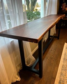 a wooden table sitting in front of a window next to a rug and white curtains