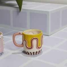 two coffee mugs sitting next to each other on a table with a plant in the background