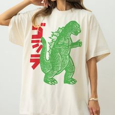 Take a step back in time with our Vintage Japanese Godzilla Art T-Shirt!  Iconic Design Meets Timeless Style: Retro Appeal: Featuring a bold, graphic print inspired by classic Godzilla movie posters, this shirt is a nod to the King of Monsters and retro Japanese art. Vibrant Artwork: The vivid green monster print against the charcoal grey tee makes a statement that's both classic and on-trend. Super-Soft Cotton: Made from high-quality cotton, this t-shirt offers all-day comfort whether you're in the city or on a movie marathon at home. Flattering Unisex Fit: Our tees are designed to look great on all shapes and sizes, available from S to XXL. Refer to our size guide for your perfect fit. Durability and Quality: Lasting Print: The durable screen-printed design stays true through washes and Graphic Printed Tees, Retro Graphic Tshirt, Retro Tee Shirts, Funky Tshirts Designs, Doodle T Shirt Design, Trending T Shirt Designs, 80s Tshirt Design, Vintage T-shirt With Screen Print For Streetwear, Japanese Graphic Shirt