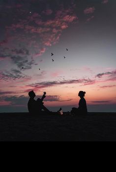 two people sitting on the ground at sunset with birds flying in the sky behind them