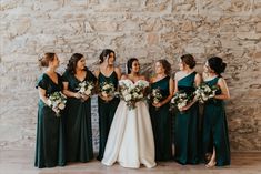 a group of women standing next to each other in front of a stone wall holding bouquets