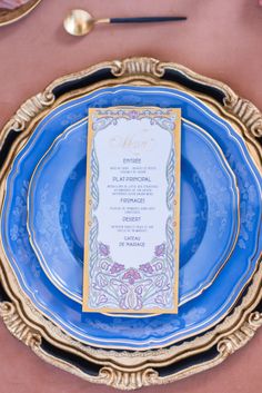 a blue and gold plate with a menu card on the front sitting on a pink table cloth