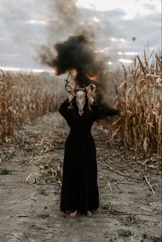 a woman in a long black dress holding up a mask over her head while standing in a corn field