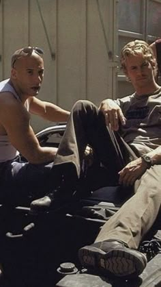 two men sitting on the hood of a car talking to each other while another man stands nearby