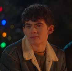 a young man sitting in front of a christmas tree wearing a jacket and scarf on his neck