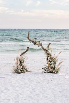 two chairs are set up on the beach for an outdoor ceremony with flowers and greenery