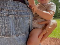 a woman holding a baby up to her chest in front of a park area with grass and trees