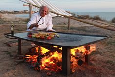 a man sitting in front of a fire pit with food on it and an umbrella over his head