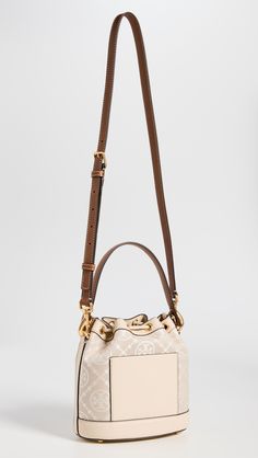 Find TORY BURCH T Monogram Bucket Bag on Editorialist. This Tory Burch bucket bag features a structured silhouette with gold-tone hardware. The bag is crafted from jacquard with a monogram pattern and leather trim. It has a drawstring top, patch interior pocket, and an optional adjustable strap. The bag is lined in suede and has a height of 8.25 inches, a length of 8.25 inches, and a depth of 4.75 inches. The strap drop is 21.75 inches and the handle drop is 4 inches. Monogram Canvas Pouch Shoulder Bag With Gold-tone Hardware, Monogram Canvas Bucket Bag With Detachable Handle, Gold Monogram Canvas Bag With Gold-tone Hardware, Designer Cream Bucket Bag, Gold Monogram Canvas Shoulder Bag With Dust Bag, Luxury Cream Bucket Bag In Pouch Shape, Luxury Cream Pouch Bucket Bag, Luxury Cream Bucket Bag, Everyday Bucket Bag In Monogram Canvas With Gold-tone Hardware