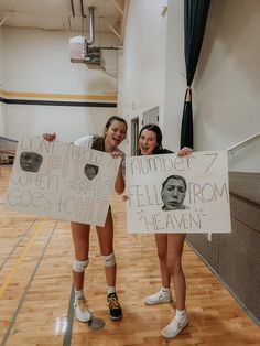 two girls holding up signs with faces on them in a gym floor, one girl is smiling and the other has her hands behind her head