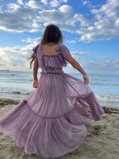 a woman in a long purple dress walking on the beach with her back to the camera