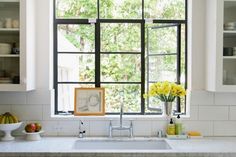 a kitchen sink under a window with yellow flowers in the vase and fruit on the counter