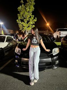 a woman standing next to two cars in a parking lot at night with her hands on the hood