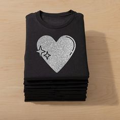 a stack of black t - shirts with silver glitter heart and stars on the front